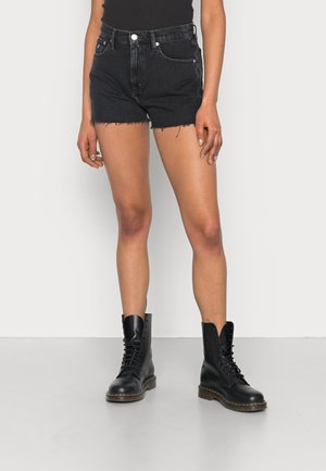 Vrouw draagt zwarte high-waisted spijkershorts met rafelige zoom, zwarte veterboots en een zwarte mouwloze top tegen een effen achtergrond.