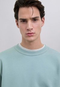 Sudadera azul clara hecha de una tela suave; cuenta con un cuello acanalado y un corte holgado. El fondo es neutro, lo que resalta la prenda.