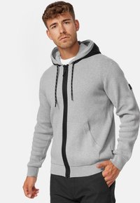 Sweat à capuche gris zippé avec une capuche noire, présentant un design texturé, des accents avec cordons de serrage et des poches avant. Poignets et ourlet côtelés.