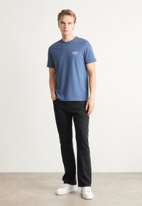 T-shirt blu in cotone con un piccolo logo bianco sul petto, abbinata a jeans neri a gamba dritta e sneaker bianche.