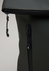 Grön vattentät ryggsäck med rulltoppsdesign, utrustad med ett sidofack med dragkedja och svarta dragkedjor. Slät textur med synlig sömnad.