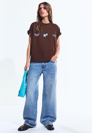 Žena stojí, má na sebe hnedé tričko s grafickým motívom so zrolovanými rukávmi, široké modré džínsy, čierne topánky a v pravej ruke drží modrú kabelku so cvokmi.