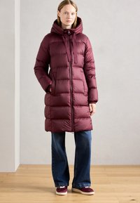 Bordeaux pufferjack met een capuchon, ritsluiting en elastische trekkoorden, voorzien van een gewatteerd ontwerp en twee zijzakken, gecombineerd met wijde jeans.