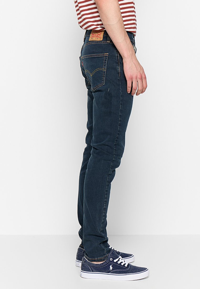 Slim-fit ciemnoniebieskie dżinsy z żółtym przeszyciem, pięć kieszeni, skórzana naszywka na tylnym pasku. W połączeniu z granatowymi sneakersami.
