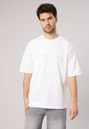 Man met donker haar die een wit oversized t-shirt draagt met de tekst "Deeluxe Original" en lichtgrijze jeans, tegen een effen achtergrond.