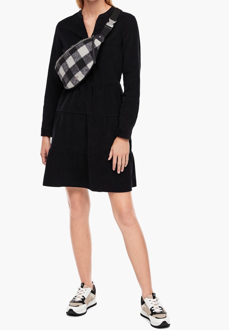 Robe en velours côtelé noire avec un design à volants, manches longues et col à revers. Associée à une banane à carreaux et des baskets.