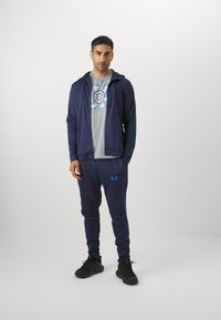 Marine treningsdrakt med blå detaljer, som inkluderer en hettegenser med glidelås og en grå T-skjorte. Sorte joggesko fullfører antrekket.