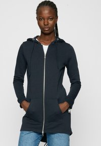 Sweatshirt zippé bleu marine en mélange de coton, doté d'une grande poche avant, d'une capuche avec cordon de serrage ajustable et de poignets en côtes. Design long.