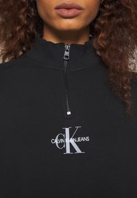 Černá zipová mikina vyrobená z měkké látky s vysokým límcem. Na přední straně je bílé logo "CALVIN KLEIN JEANS" a malý detail v podobě stříbrného zipu.