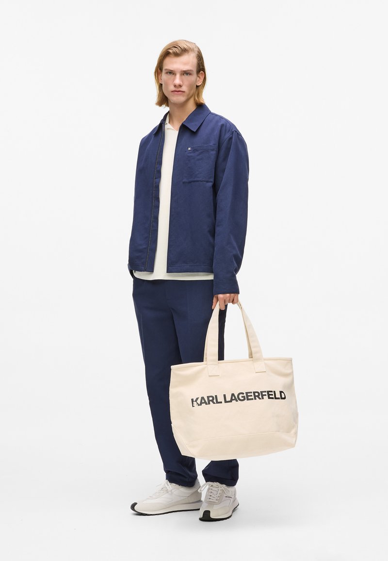 Veste-chemise bleu marine avec une poche, T-shirt beige, pantalon assorti et sac fourre-tout beige avec le texte noir "KARL LAGERFELD". Chaussures décontractées.