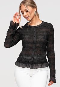 Femme portant un chemisier noir transparent à manches longues avec des volants et des sequins, associée à un pantalon blanc, posant devant un fond clair.