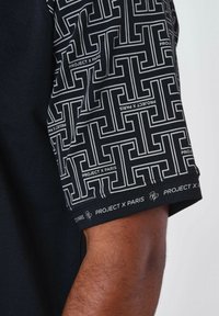Camiseta negra con mangas cortas estampadas que presentan un diseño geométrico en blanco. "PROJECT X PARIS" impreso en la manga y el dobladillo.