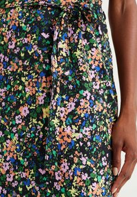 Close-up of a hand resting beside a waist with a tied belt on a multicolored floral print dress with green, orange, blue, and pink flowers.