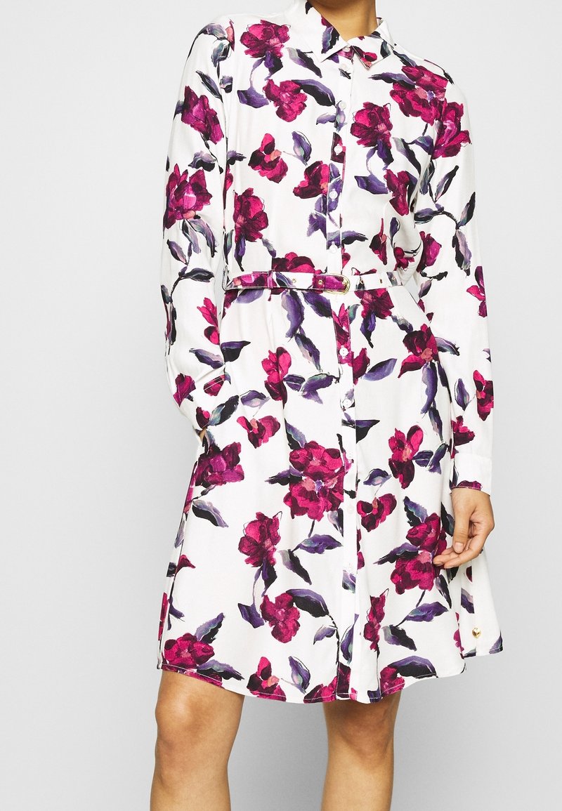 Donna che indossa un vestito camicia bianco con maniche lunghe, stampa floreale rosa e viola, e una cintura abbinata in vita.