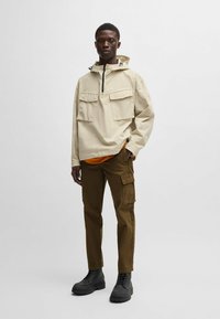Beige hoodie met twee voorkruizen, zwarte rits en oranje binnenlaag, gecombineerd met bruine cargo-broek en zwarte veterschoenen.