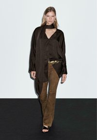 Top in raso marrone con dettaglio sciarpa, abbinato a pantaloni svasati in suede marrone e sandali neri con cinturini, su sfondo neutro.