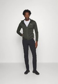 Temno zelena kapucar s kapuco, z zadrgo, združena z oprijetimi črnimi hlačami z žepi. Model stoji z eno roko na boku, ozadje je minimalno.