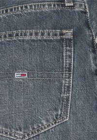 El bolsillo trasero de tela de mezclilla en azul oscuro con costuras amarillas. Presenta un pequeño logotipo con franjas en rojo, blanco y azul encima del bolsillo.