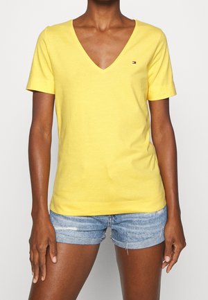 Żółta, krótko rękawowa koszulka z dekoltem w serek, wykonana z bawełny, z małym logo na piersi; zestawiona z postrzępionymi szortami dżinsowymi. Gładka faktura.