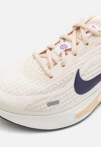 Zapatilla de deporte color beige claro con parte superior de malla texturizada, logotipo de Nike púrpura, suela acolchada y acentos en color crema. Presenta cordones y una pequeña etiqueta de marca.