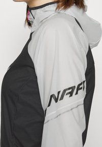 Veste légère noire et grise avec capuche, poignets élastiques et logo latéral proéminent. Tissu texturé avec accents en mesh.