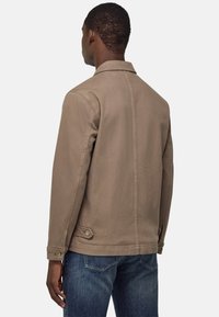 Giacca di cotone marrone con colletto, cuciture sulle spalle e dettagli con bottoni sui polsini, abbinata a jeans in denim blu.