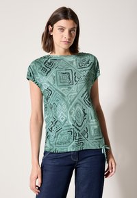 Haut à manches courtes vert à motifs présentant des designs géométriques et un détail à nouer à l'ourlet, fabriqué en tissu léger au toucher doux.