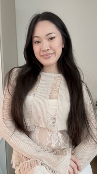 Jeune femme aux longs cheveux foncés portant un pull beige en maille usée par-dessus un top en dentelle blanche, souriante, debout dans un cadre intérieur neutre.