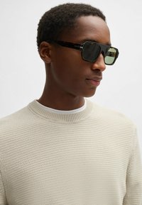 Occhiali da sole con lenti scure in una montatura esagonale effetto tartaruga. Il modello indossa un maglione testurizzato color crema sopra una camicia bianca.