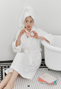 Accappatoio bianco in morbido tessuto e asciugamano, vasetto trasparente rosa di prodotti per la cura della pelle tenuto in mano, posati su un pavimento a piastrelle bianche e nere accanto a una vasca da bagno bianca.