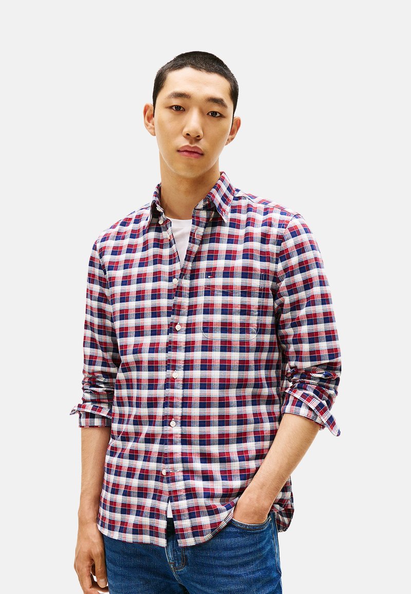 Camisa de cuadros abotonada en rojo, azul marino y blanco; con mangas largas, cuello y bolsillo en el pecho izquierdo; se lleva sobre una camiseta blanca.