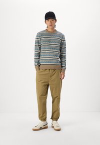 Pulover tricotat cu un model geometric în nuanțe de albastru, maro și crem, asortat cu pantaloni kaki și adidași albi, purtat cu o șapcă neagră.