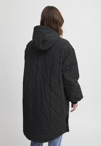 Sort, kviltet lang jakke med hætte, har diamantmønster og snap-lukninger i siden. Stoffet ser glat ud med en mat finish.