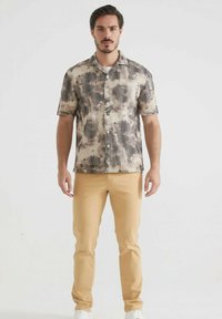 Chemise à manches courtes avec un motif abstrait gris et marron. Pantalon beige, baskets blanches. Coupe moderne, design à col boutonné, boutons visibles.