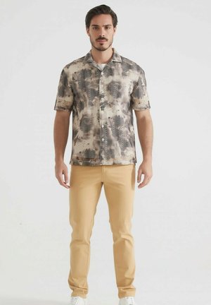 Chemise à manches courtes avec un motif abstrait gris et marron. Pantalon beige, baskets blanches. Coupe moderne, design à col boutonné, boutons visibles.