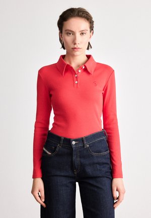Polo vermelho de mangas longas com textura canelada, com abertura de botões e gola, combinado com jeans de ganga azul escuro com costuras visíveis.