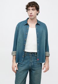 Πουκάμισο από τζιν σε μεσαίο μπλε χρώμα με κουμπιά στο μπροστινό μέρος, μανίκια που τυλίγονται και αντίθετες γκρι μανσέτες. Φορεμένο πάνω από λευκό αμάνικο τοπ και συνδυασμένο με μπλε τζιν παντελόνια.