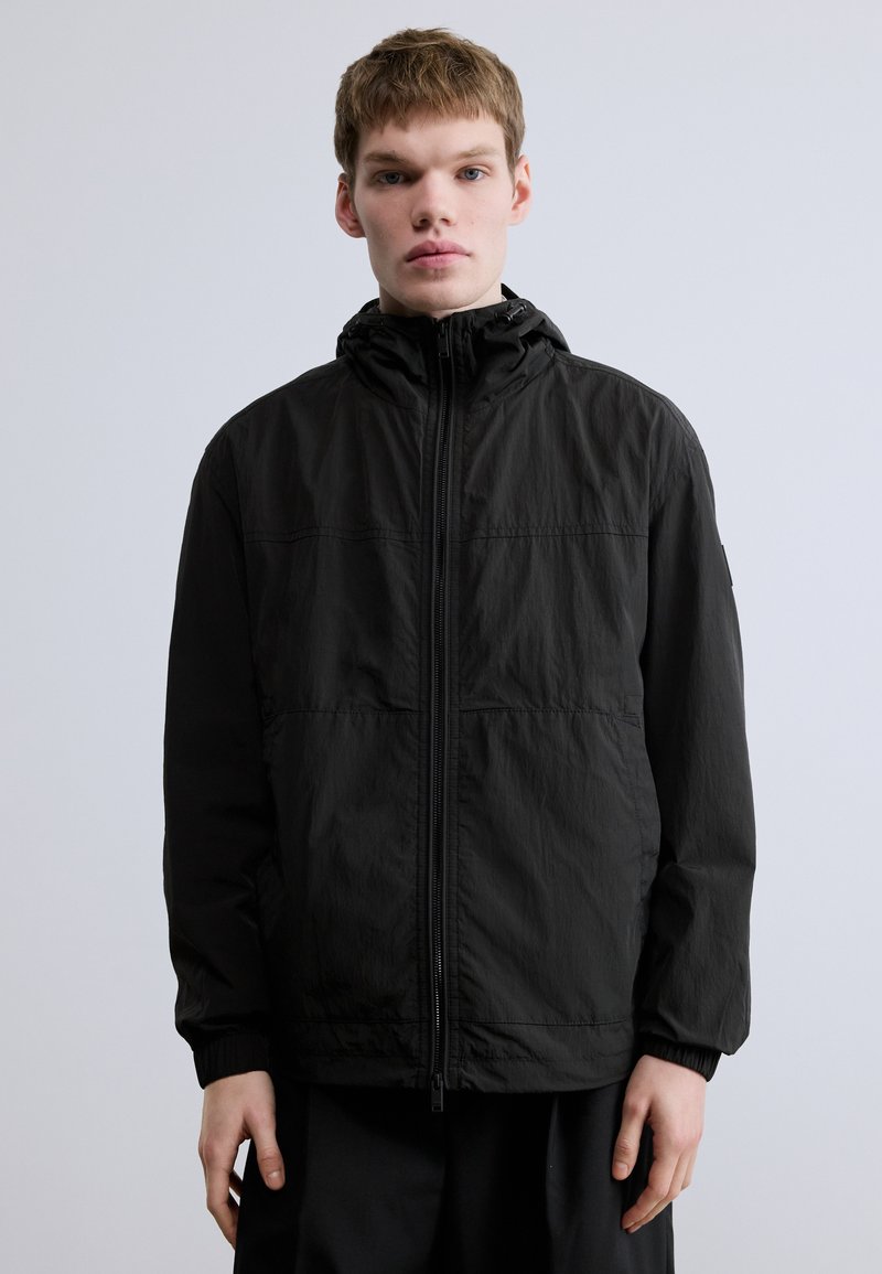 Must tuulekindel jakk kõrge kraega, esikäepidemega lukuga, elastsete varrukaservadega ja tekstuurse viimistlusega. Minimalistlik disain ilma nähtavate logodeta.