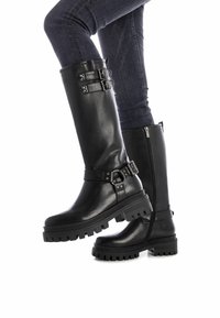 Botas de cuero negro hasta la rodilla con suelas gruesas, accents de hebilla y cremalleras laterales, combinadas con jeans oscuros. Acabado texturizado y diseño robusto.