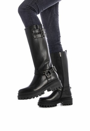 Stivali alti neri in pelle con suole spesse, dettagli con fibbie e cerniere laterali, abbinati a jeans scuri. Finitura testurizzata e design robusto.