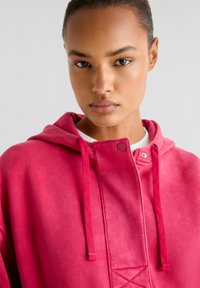 Hoodie fuchsia avec fermeture à snaps à l'avant et cordons de serrage. Caractéristiques d'une texture douce et d'une finition cousue au niveau de la poitrine.