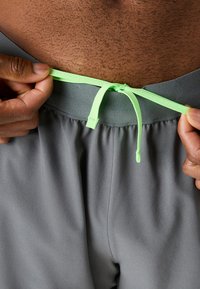 Grå træningsshorts med en glat tekstur, der har en elastisk talje og en lys grøn snoretræk. Nærbillede af snoretrækket, der bliver justeret.