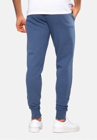 Modré tepláky s úzkým střihem, žebrovanými manžetami a bočními kapsami, vyrobené z měkké látky, kombinované s bílými teniskami.