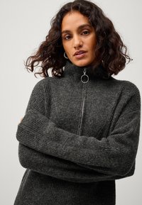 Grijze zip-up trui met hoge kraag, geribbelde textuur en lange mouwen. Voorzien van een zilveren ringvormige trekker aan de rits. Zachte stof.