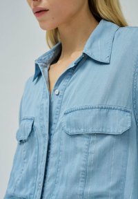 Camisa de mezclilla de color azul claro con rayas verticales blancas, cuello puntiagudo y dos bolsillos en el pecho. Presenta un cierre frontal con botones y una textura suave.