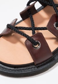 Brown leather sandal with black braided straps, metal accents, and a textured black rubber sole. Smooth tan footbed visible.