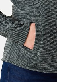 Texturerad grå tröja med ett vertikalt randmönster och sidoficka. Materialet verkar mjukt med en subtil lyster. Närliggande blått tyg synligt.