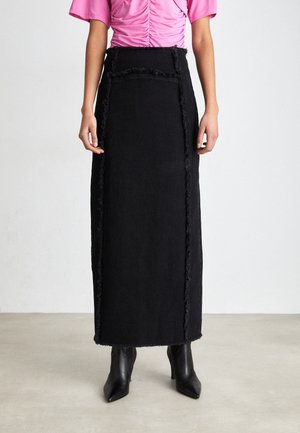 Femme portant une jupe longue noire taille haute avec des coutures effilochées, des bottines noires à talons et bouts pointus, et un haut rose à manches courtes froncées.