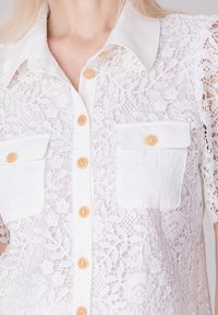 Chemisier en dentelle blanche à boutons, doté d'un col, de deux poches avant avec des accents de boutons, et d'un tissu transparent et texturé.