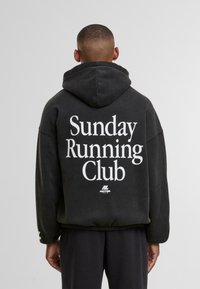 Uomo che indossa un hoodie nero con la scritta "Sunday Running Club" in bianco, in piedi di spalle contro uno sfondo grigio uniforme.